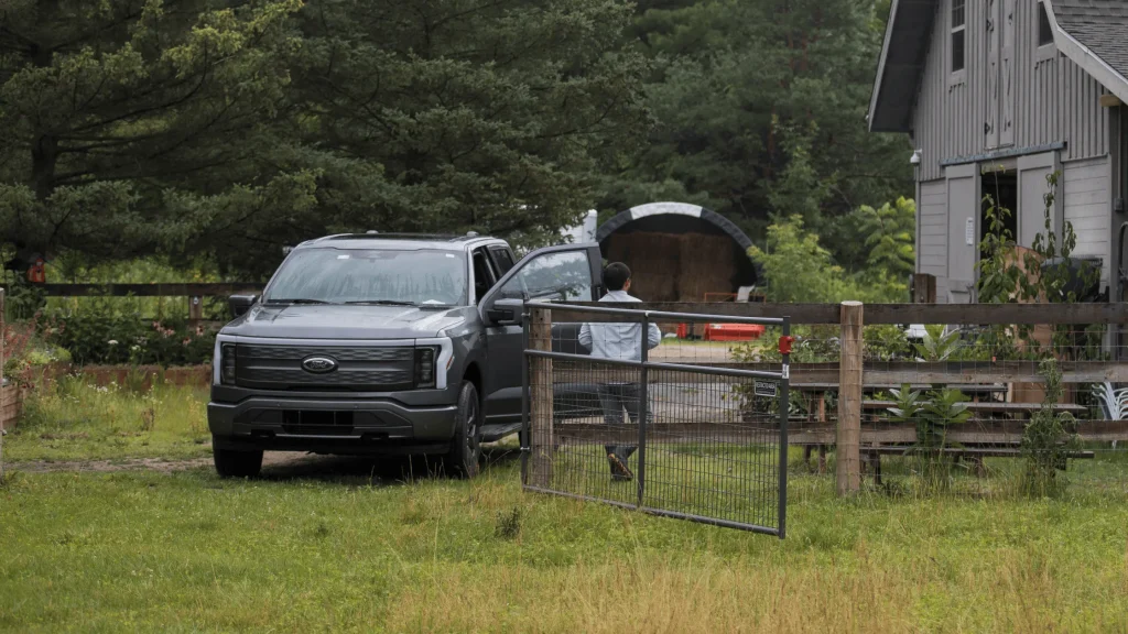 2024 Ford F-150 Lightning parked on grass beside a barn and garden with a person opening the door, illustrating rural utility.