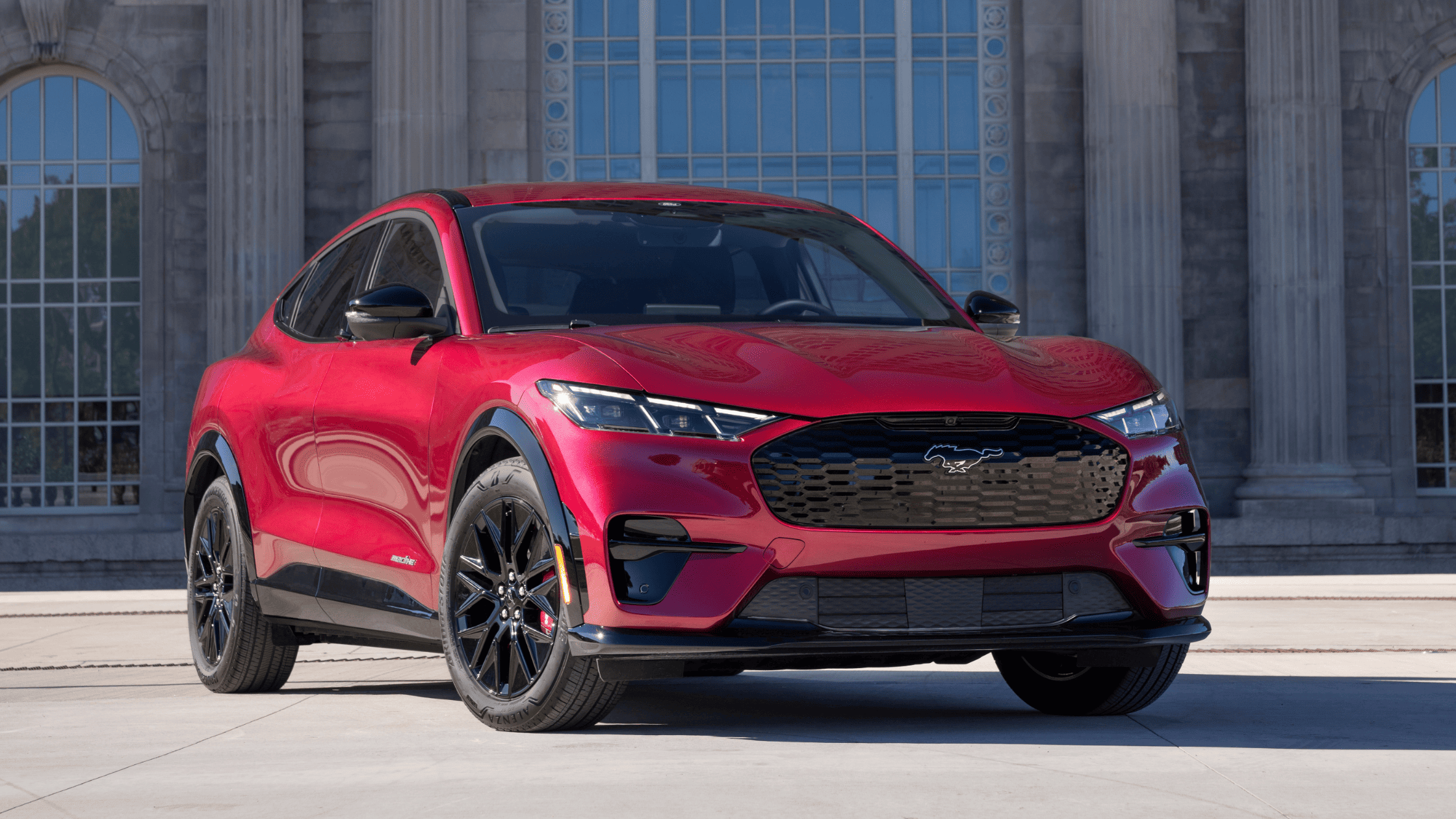 Front angle of a red 2025 Ford Mustang Mach-E® parked in front of a historic building, showcasing its EV design, Mustang heritage, and blacked-out grille.