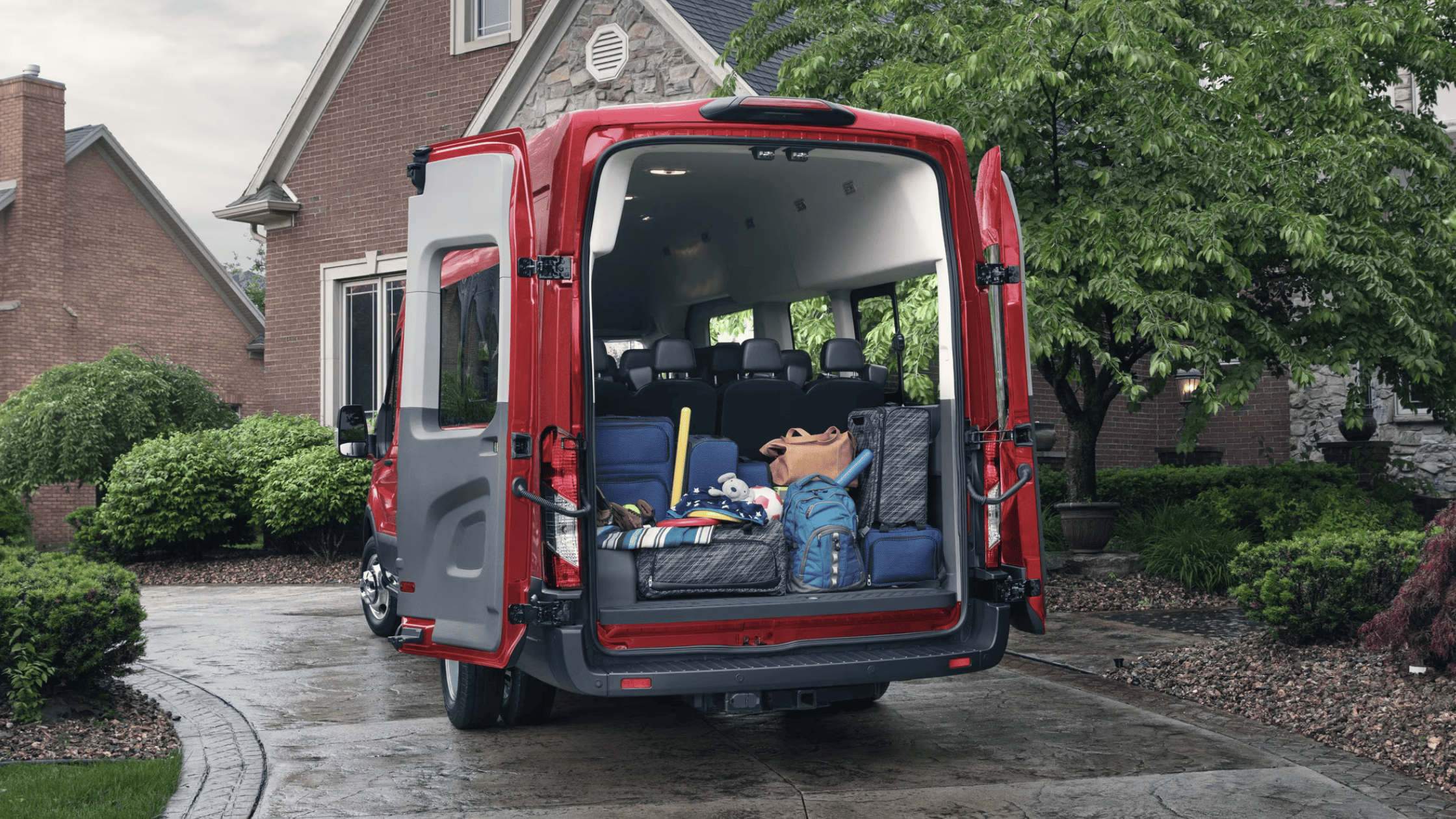 Rear cargo view of a 2025 Ford Transit® parked in a driveway, loaded with suitcases, sports gear, and backpacks for a family outing.