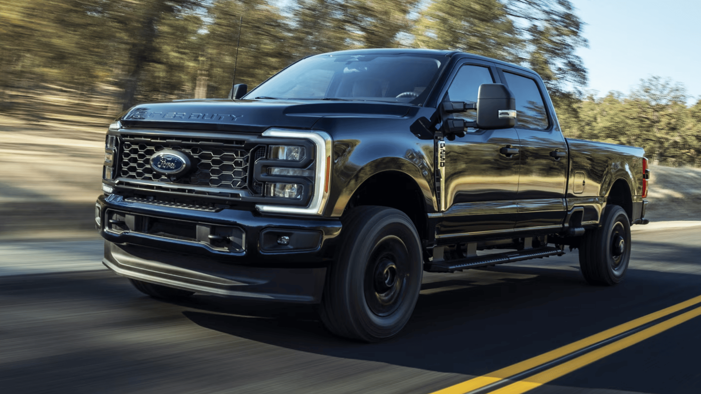 A black 2025 Ford Super Duty F-250 driving on a scenic road, highlighting its bold grille, LED headlights, and rugged exterior built for performance.
