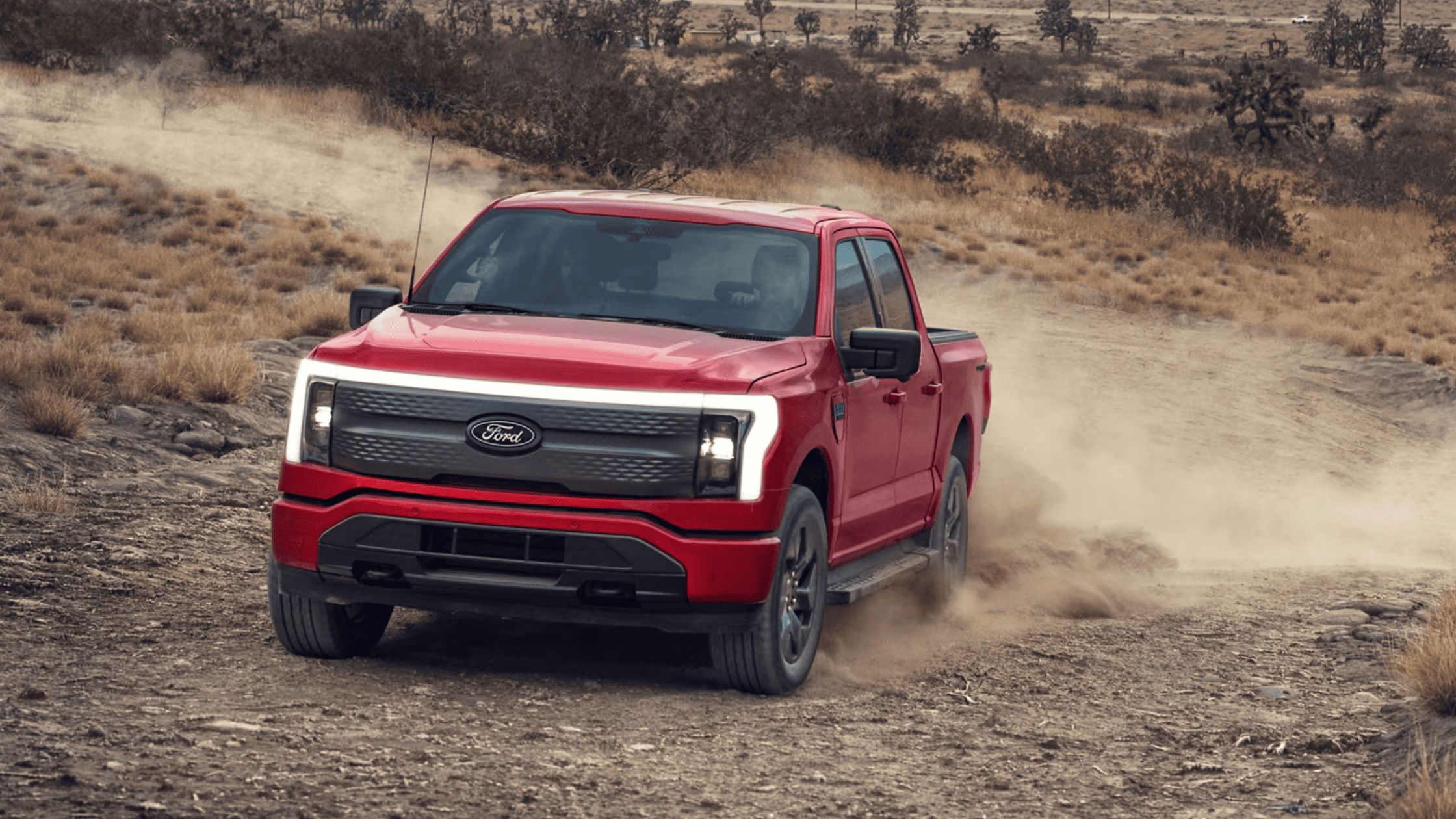A red 2024 Ford F-150® Lightning driving on a rugged off-road trail, kicking up dust, highlighting its electric power and all-terrain capability.