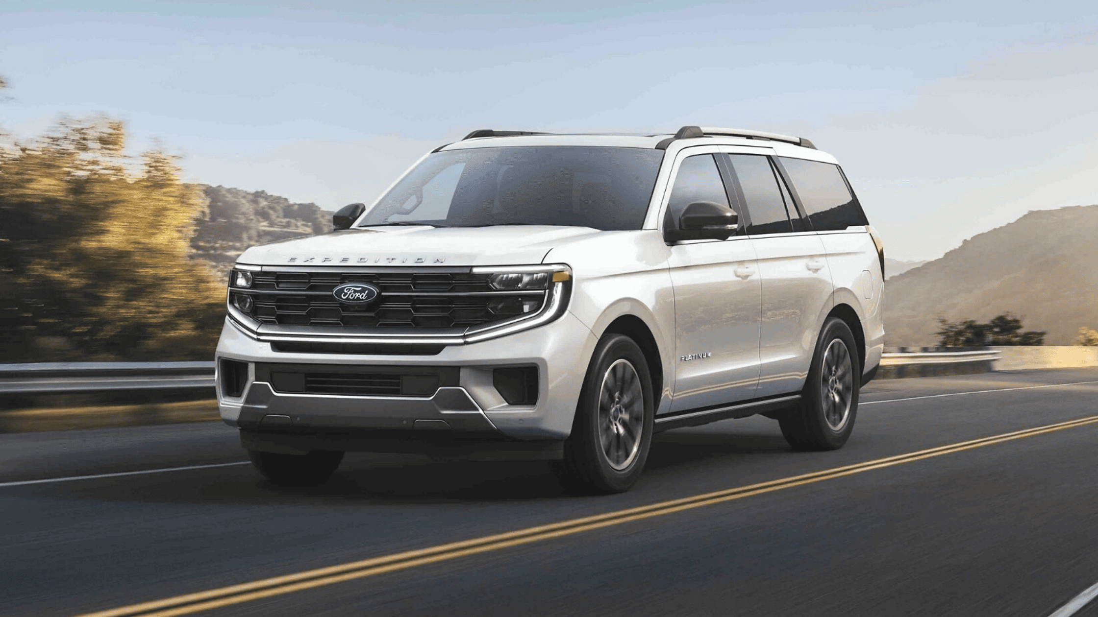 A white 2025 Ford Expedition Platinum SUV driving on a scenic highway, showcasing its bold front grille and modern design.