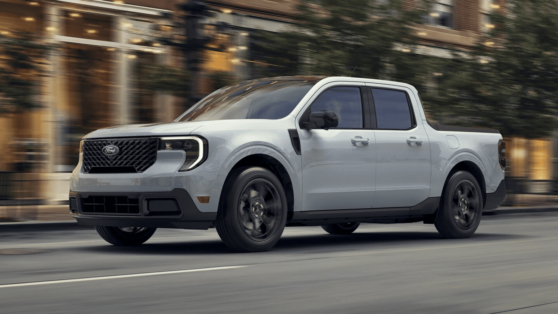 A silver 2025 Ford Maverick driving on a city street, showcasing its bold front grille, LED headlights, and stylish exterior design.