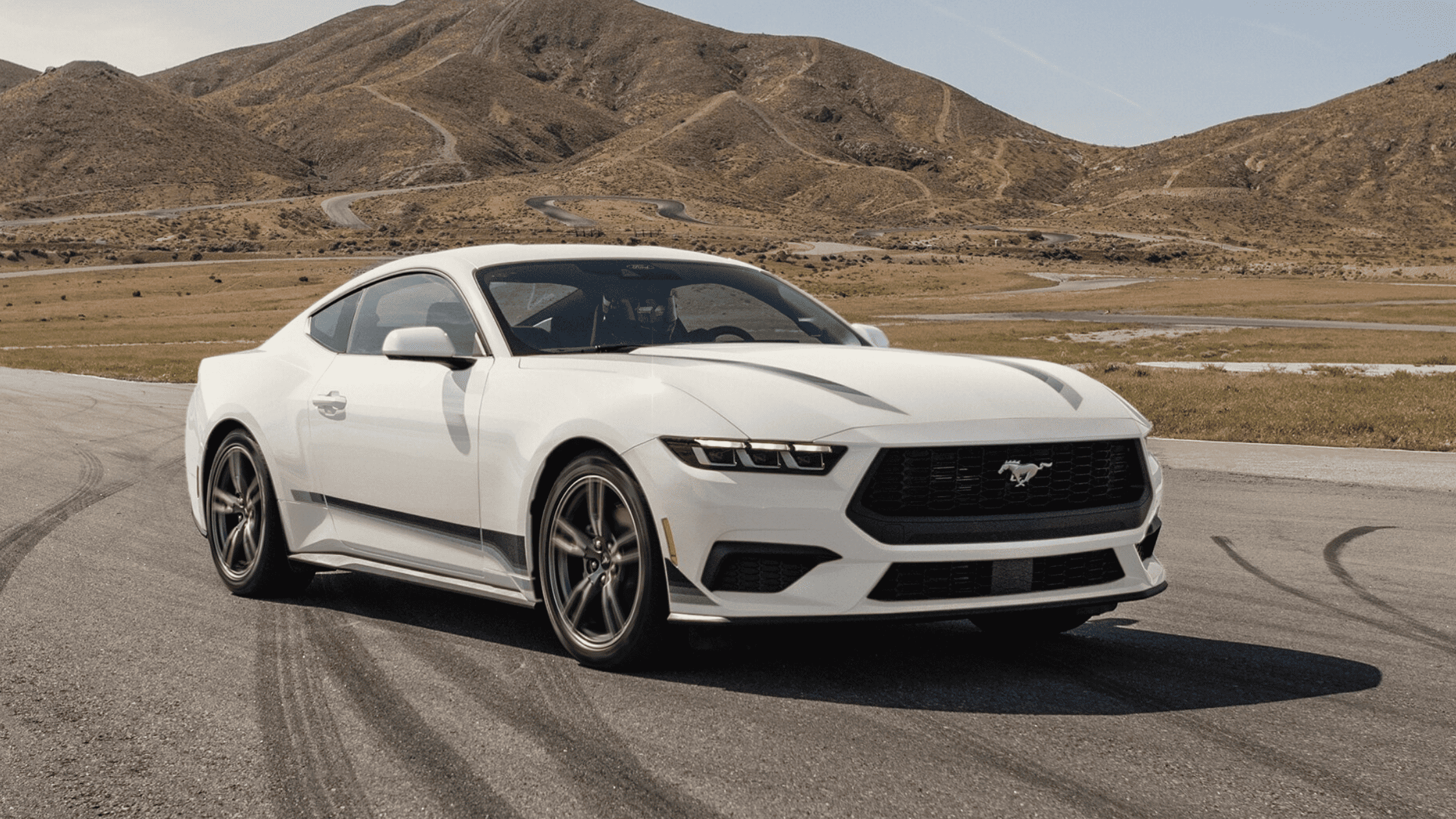 A white 2025 Ford Mustang parked on a scenic mountain road, highlighting its sleek aerodynamic design, aggressive front grille, and performance-driven styling.
